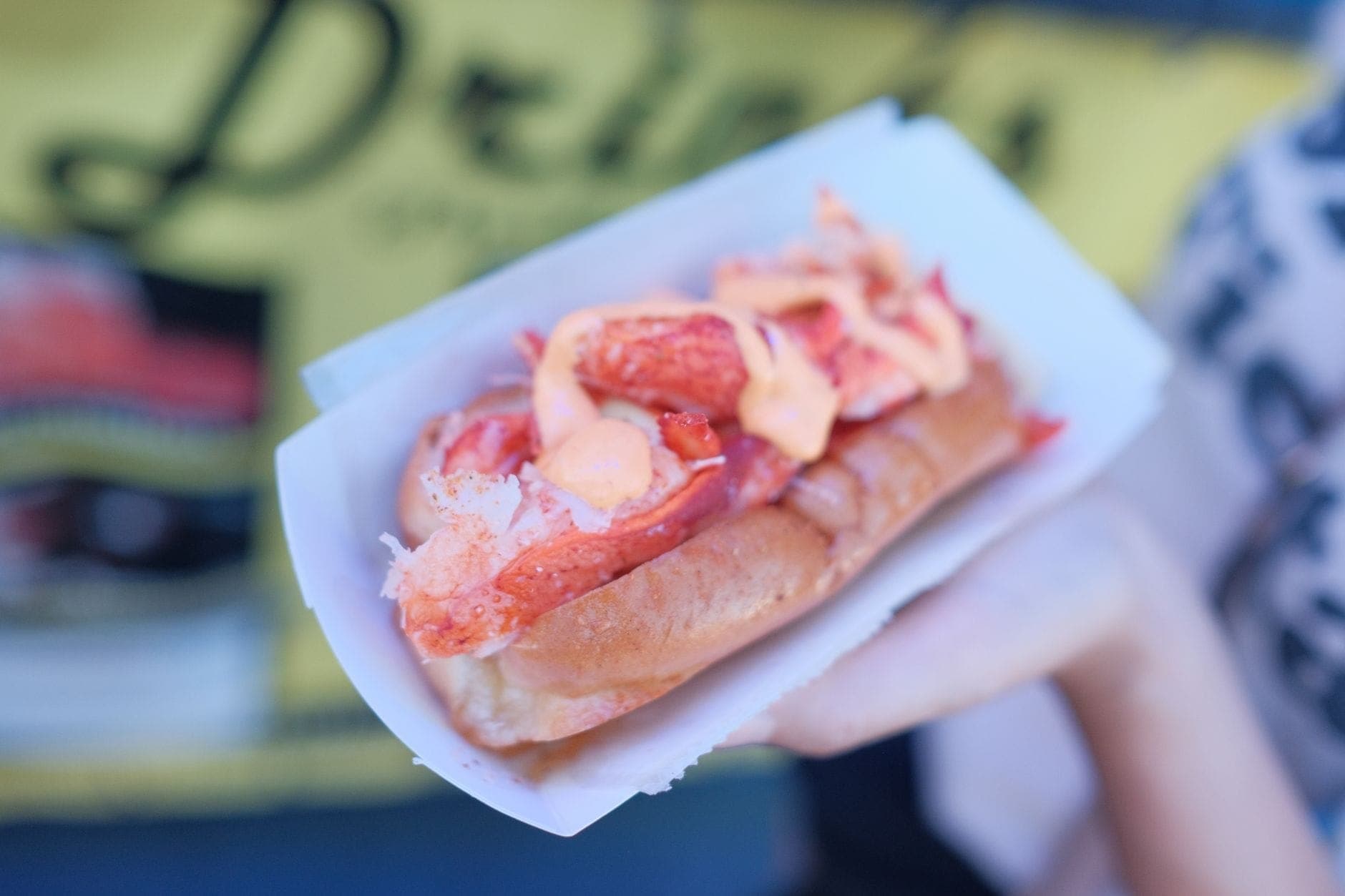 Une main tient un lobster roll classique de style Nouvelle-Angleterre dans un plateau en papier, garni de chair de homard frais sur un pain beurré.