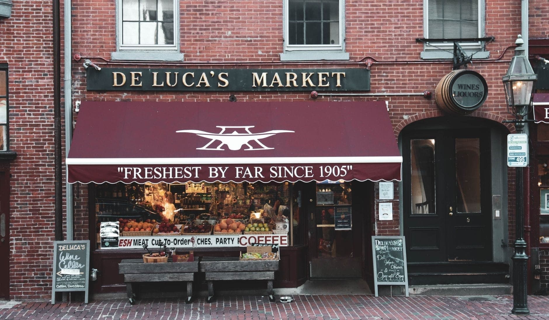 Devanture historique du De Luca's Market à Boston, avec son auvent rouge, ses produits frais en vitrine et une atmosphère de quartier chaleureuse dans une rue pavée de briques.