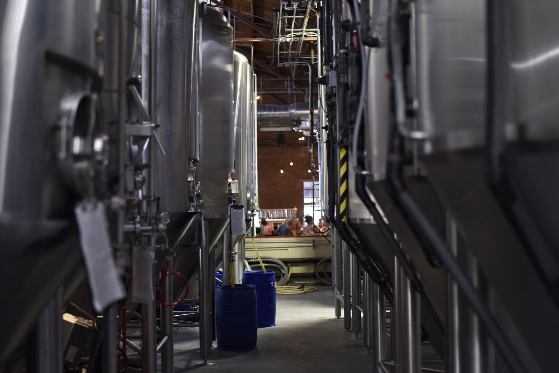 De grands cuves en acier pour la brasserie longent un couloir avec un aperçu de personnes assises à des tables en arrière-plan, suggérant une brasserie active.