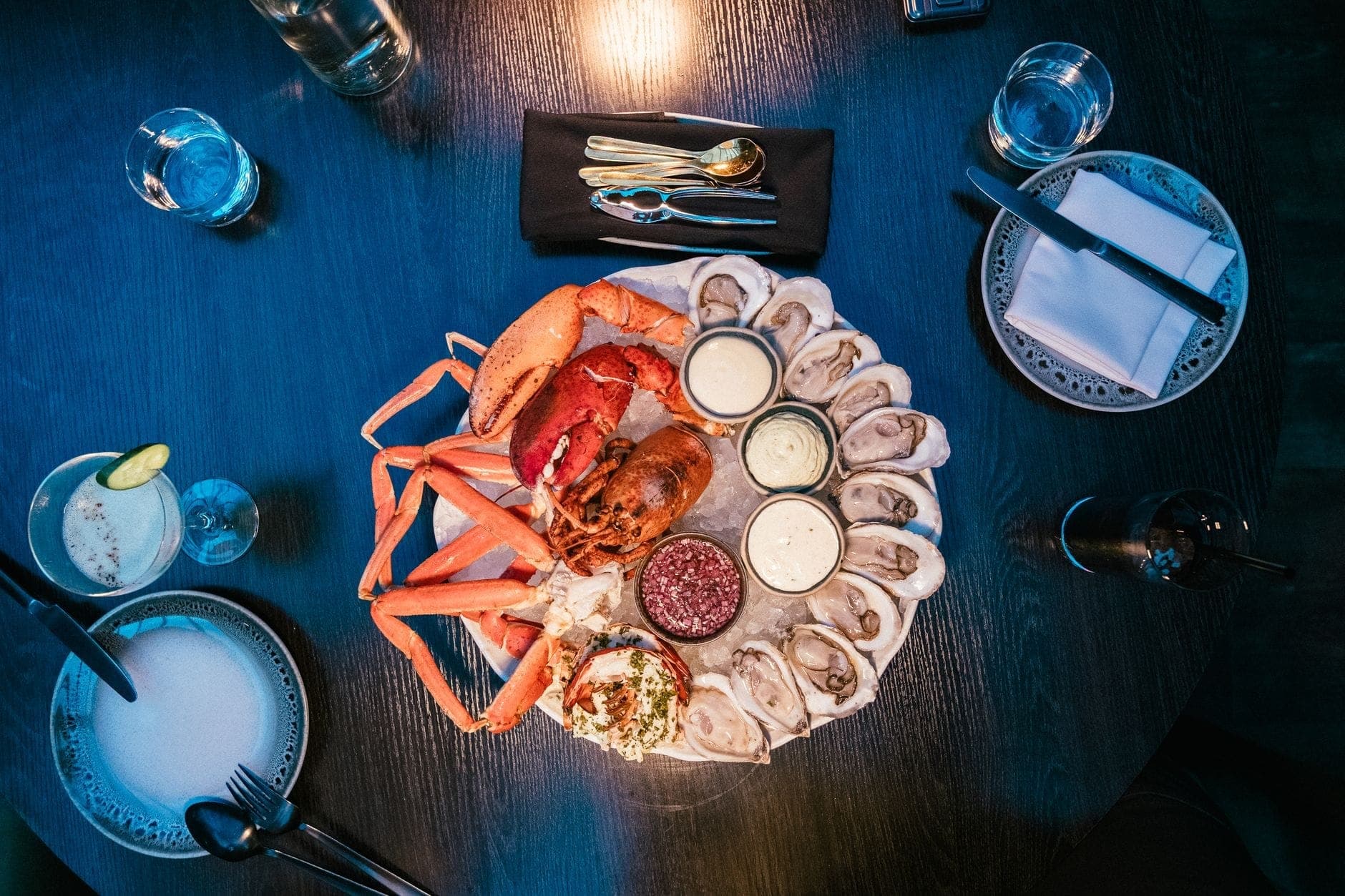 Plateau d'huîtres, de homard et de crabe sur glace avec des sauces sur une table de restaurant avec boissons et couverts.
