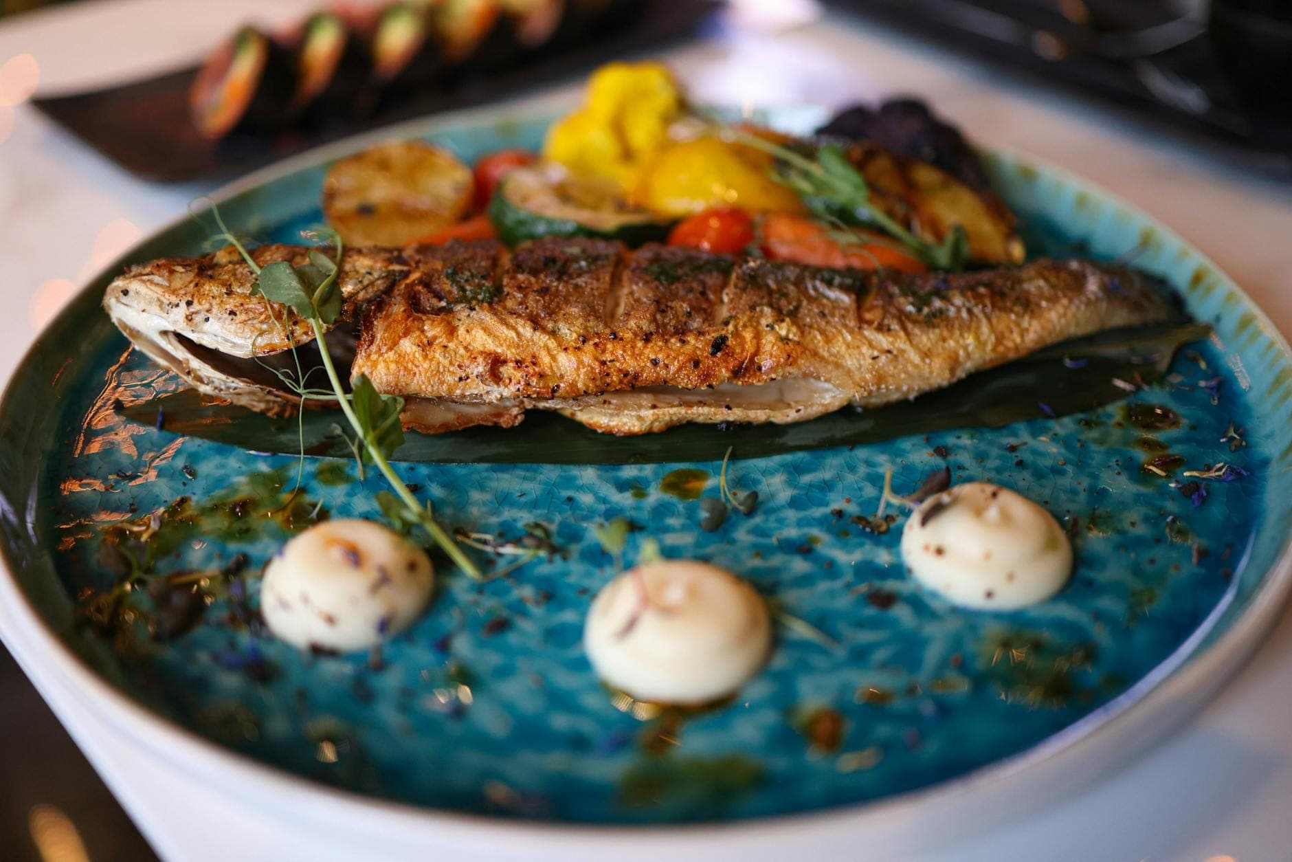 Un poisson entier grillé servi sur une assiette en céramique bleue avec une garniture de légumes, joliment présenté dans un cadre de restaurant.