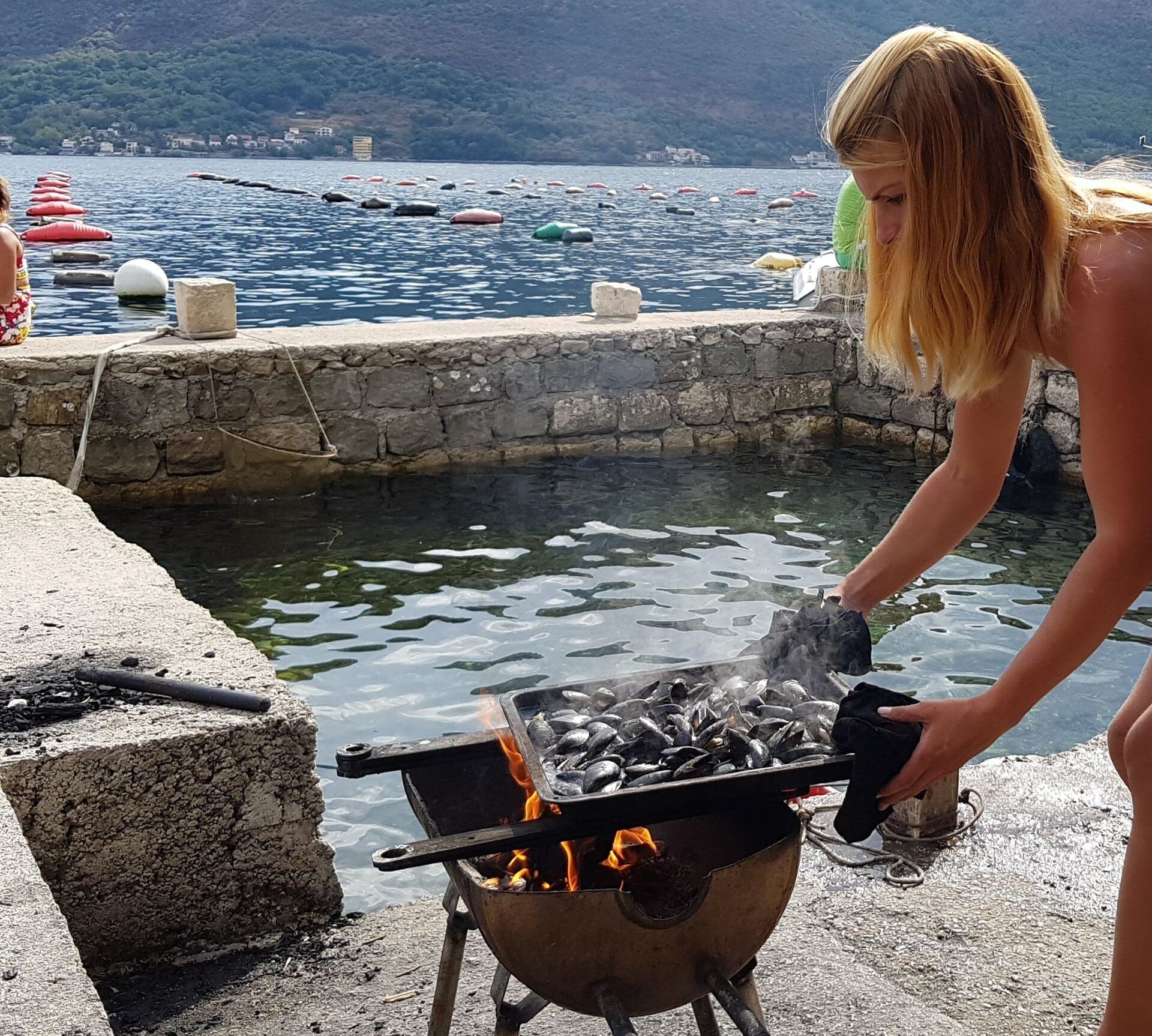 Une femme fait cuire des moules fraîches sur une flamme vive au bord de l'eau à Kotor, avec la baie de Boka et les montagnes en arrière-plan.