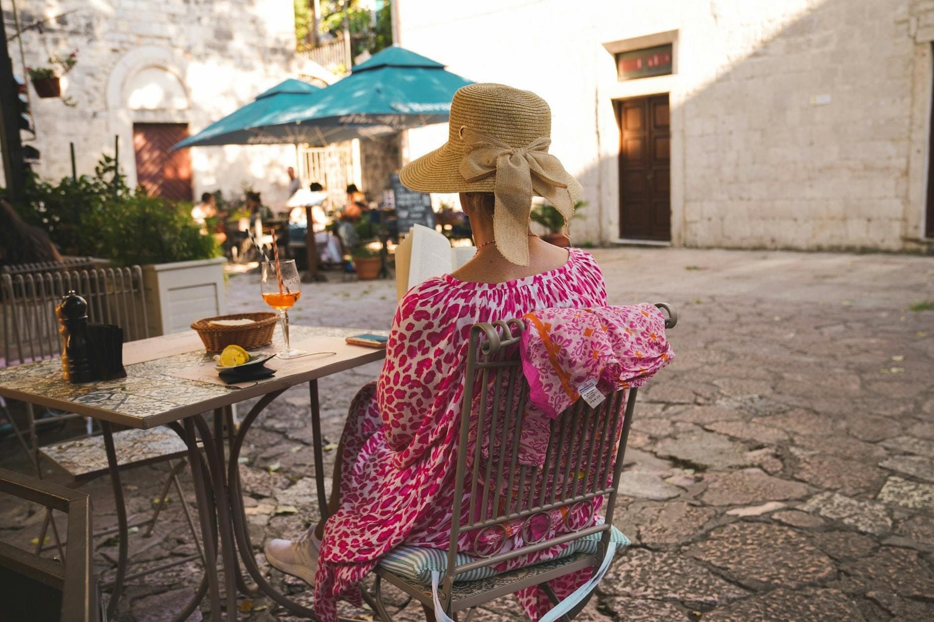 Femme en chapeau de paille assise à une table de café en terrasse à Kotor avec une boisson et un Aperol Spritz visibles, fond de place en pierre.
