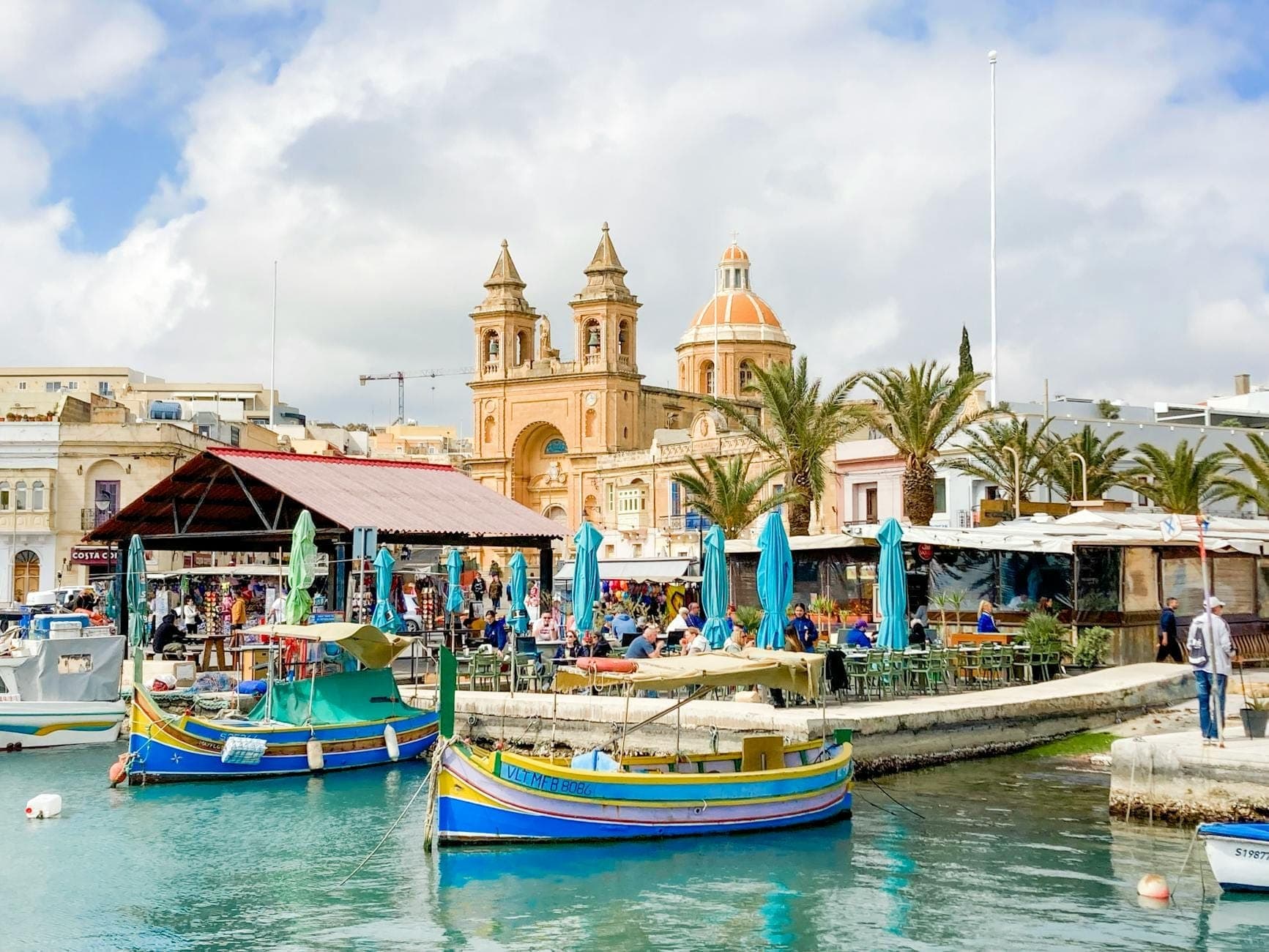 Animado paseo marítimo maltés con coloridos botes de pesca, restaurantes al aire libre y una prominente iglesia, que captura una auténtica escena de mercado y gastronomía.