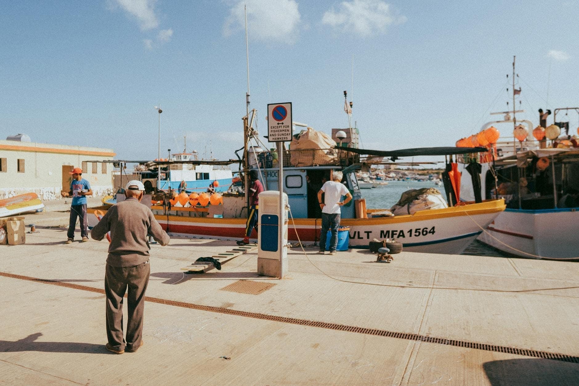 Pescadores en un puerto maltés preparando botes, con aparejos de pesca y muelles visibles bajo cielos soleados.