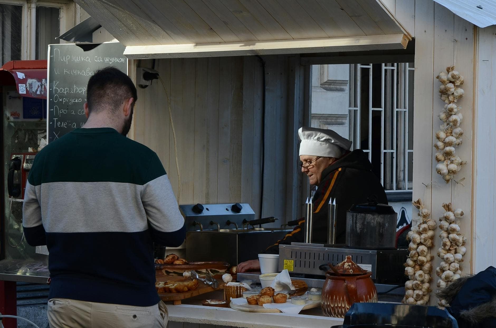 Un hombre comprando pasteles en un pequeño puesto de comida callejera donde una persona con gorro de chef está sirviendo. Los pasteles están expuestos al frente.