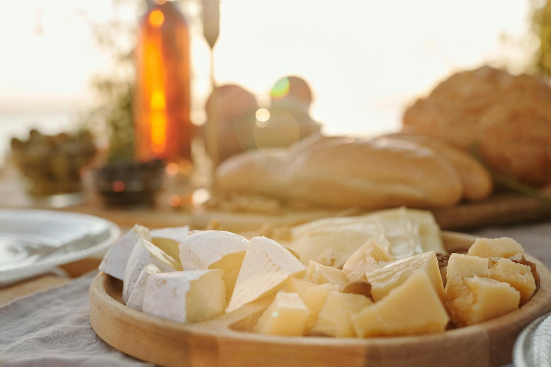 Tabla de madera con quesos variados, pan, aceitunas y botellas de vino sobre una mesa al aire libre bajo el sol, evocando un ambiente mediterráneo