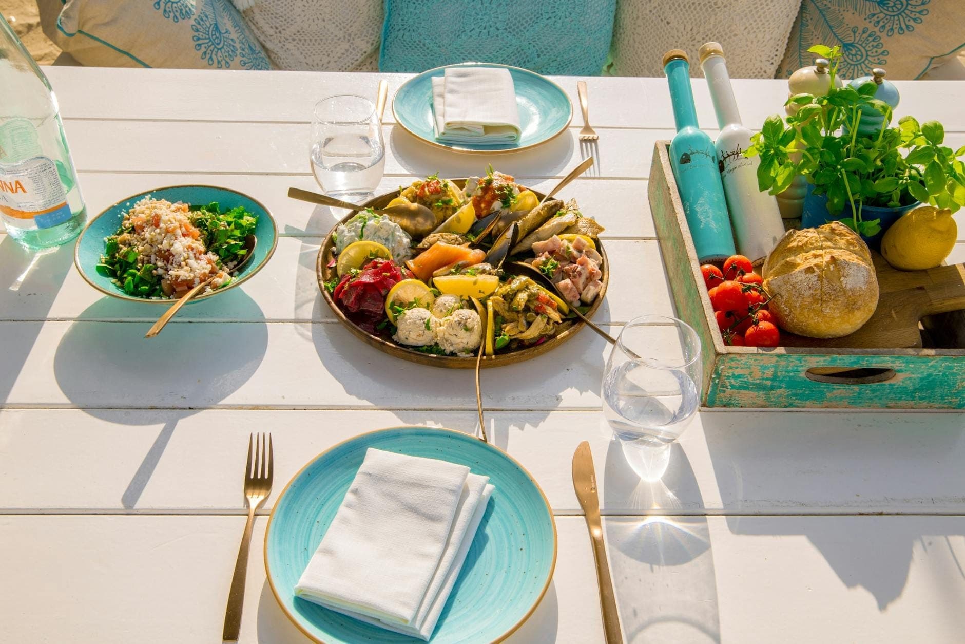 Buntes griechisches Mezze-Tablett, Salat, Brot, Kräuter und Getränke auf einem sonnenbeschienenen weißen Tisch in Mykonos, mit türkisfarbenen Tellern und gemütlichen Kissen.
