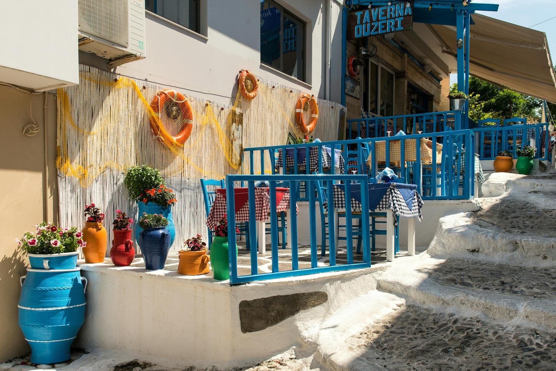 Bunte Außensitzplätze einer griechischen Taverne mit blauen Geländern, karierten Tischdecken und leuchtenden Blumentöpfen auf klassischen weiß gekalkten Stufen.