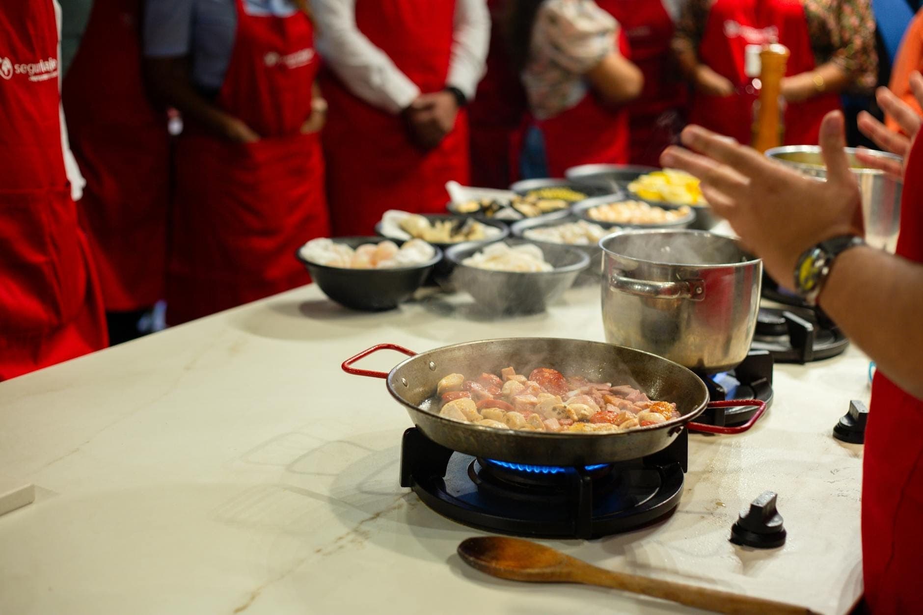Scena di corso di cucina con più persone in grembiuli rossi che osservano qualcuno preparare cibo tradizionale in una padella, illustrando tradizioni culinarie condivise.