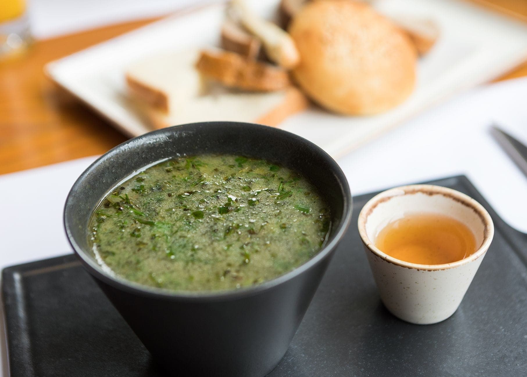 Un tazón de sopa verde con hierbas junto a una pequeña taza, con pan al fondo en una bandeja.