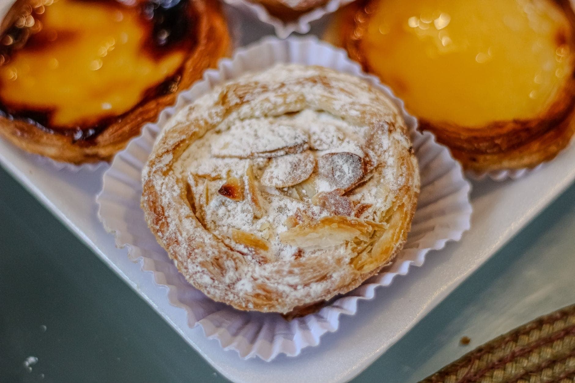 Primer plano de un pastel jesuíta cubierto con azúcar en polvo y láminas de almendra, con pastéis de nata cerca en una caja de panadería.