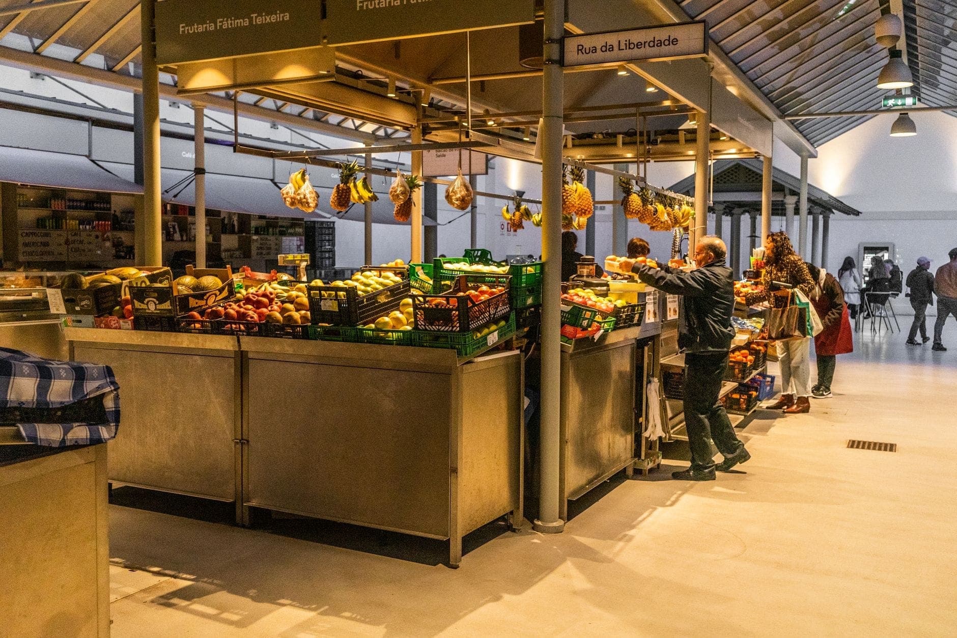 Personas comprando en puestos de mercado interior llenos de productos frescos bajo un techo de metal y cristal en Oporto, Portugal.