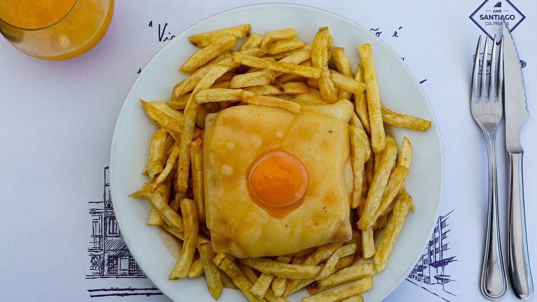Un plato de francesinha cubierta con un huevo frito y rodeada de patatas fritas, servida en un plato blanco con cubiertos y bebida naranja.