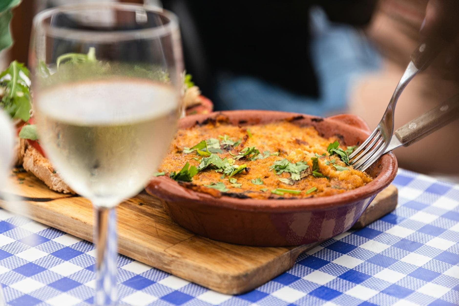 Plat grec traditionnel cuit au four dans une terrine en terre cuite rustique, garni d'herbes fraîches, servi avec un verre de vin blanc sur une nappe à carreaux bleus.