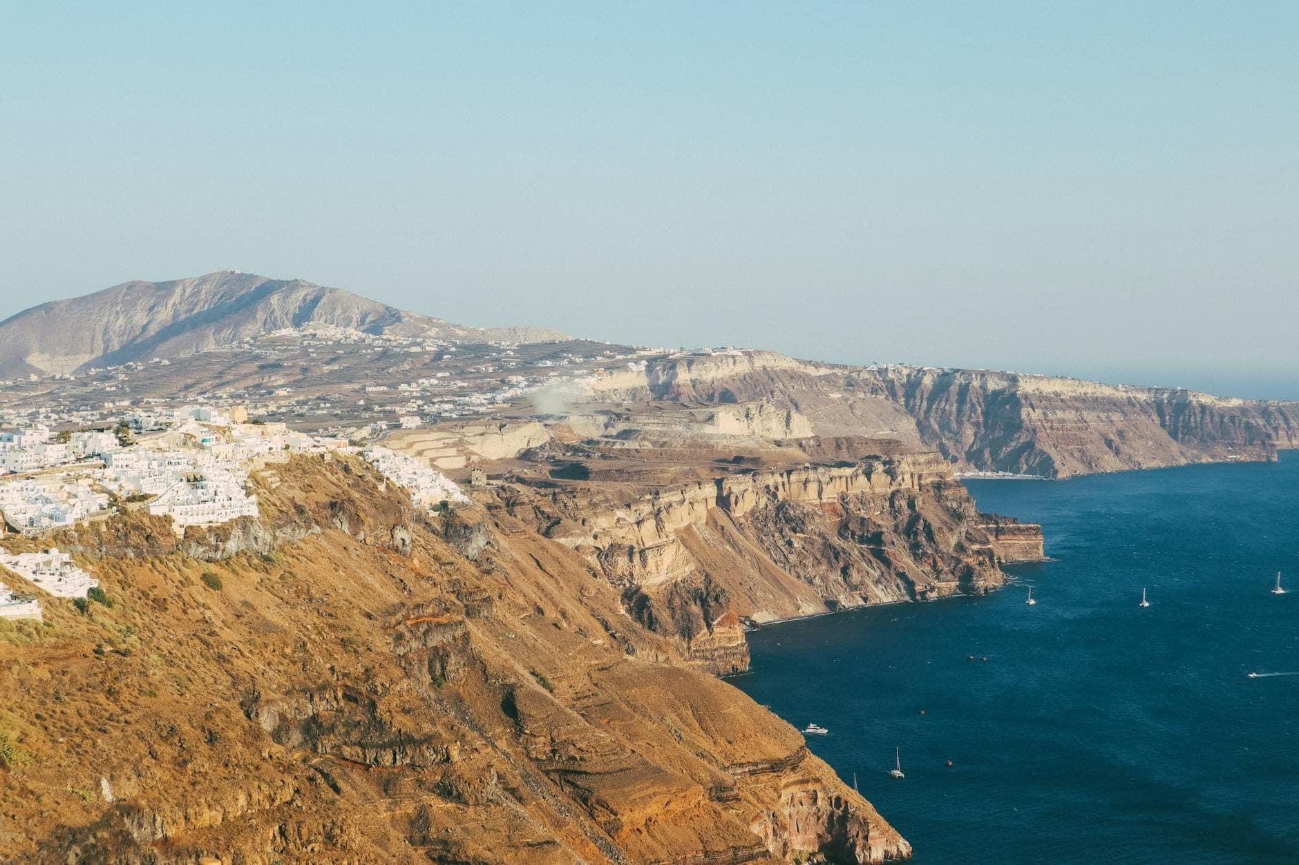 Aerial view of Santorini’s rugged volcanic cliffs with whitewashed towns perched on the edge and deep blue sea below, on a clear day.