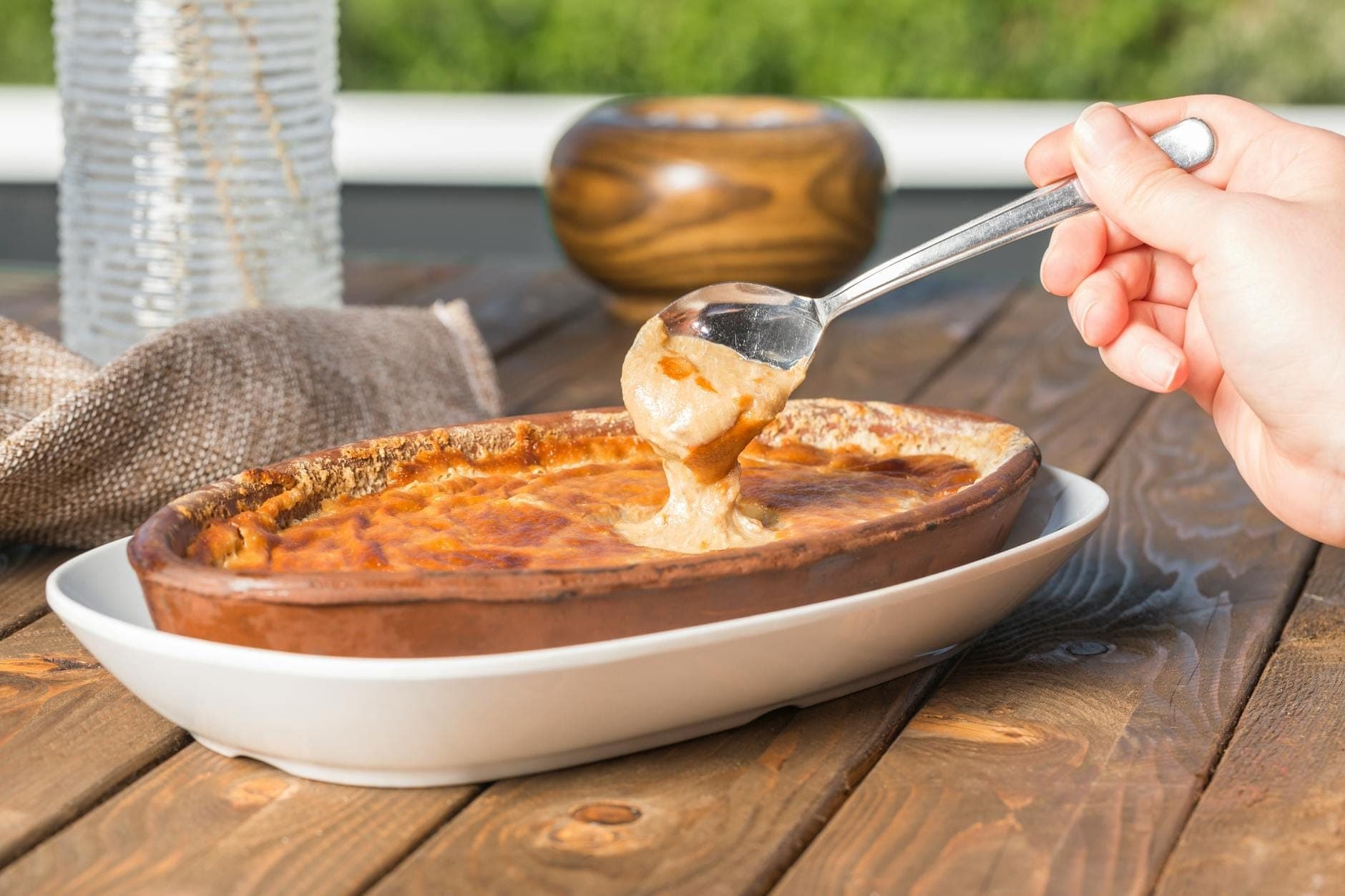 Hand scooping creamy yellow dip from oval clay dish, likely Santorini fava, on a wooden table outdoors.