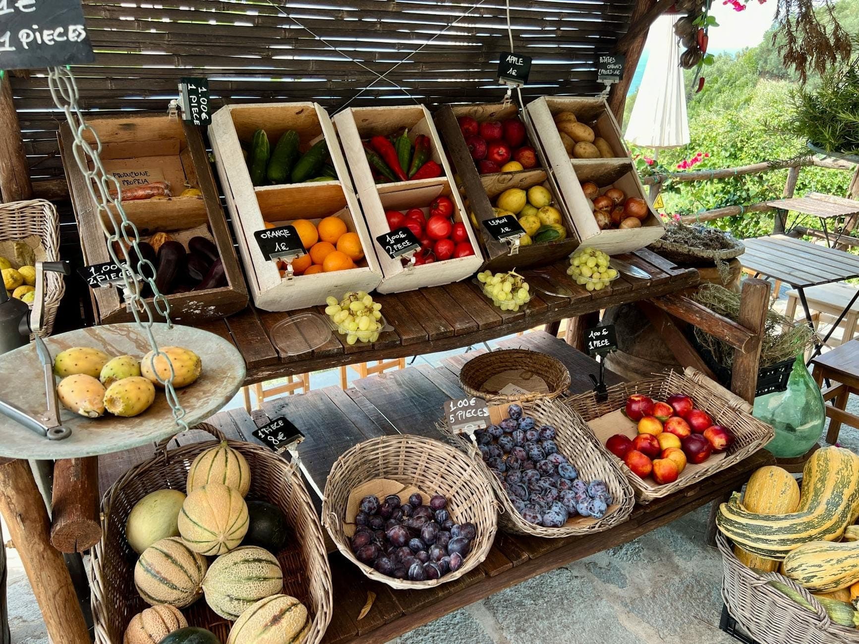 Outdoor market stand in Santorini with baskets and boxes of seasonal local produce including melons, plums, tomatoes, cucumbers, and grapes.