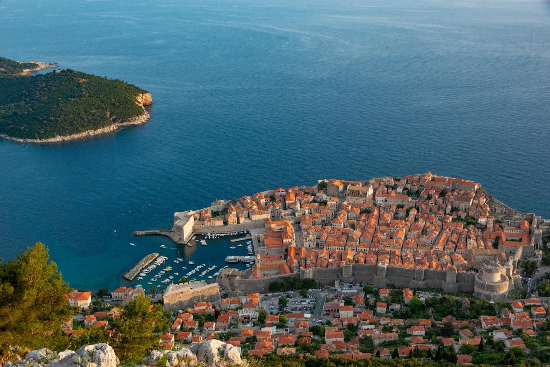 Vue aérienne de la vieille ville de Dubrovnik sur le continent avec l'île de Lokrum à proximité, séparées par une mer bleue sous un ciel dégagé.