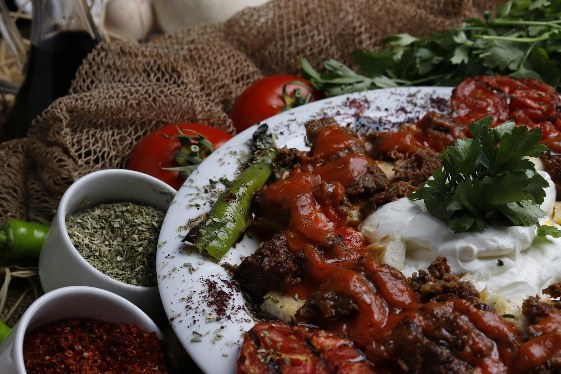Gros plan d'un plat turc avec de la viande grillée, de la tomate, du poivron, du yaourt et des herbes dans une assiette entourée d'épices et de légumes frais.