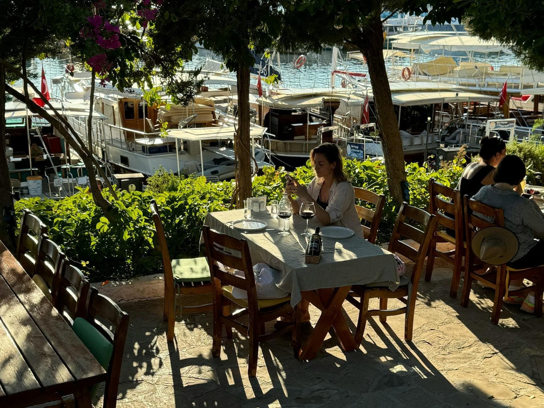 Terrasse de restaurant en plein air donnant sur la marina d'Antalya, avec des tables en bois, des arbres ombragés, des personnes qui dînent et des yachts blancs amarrés en arrière-plan.