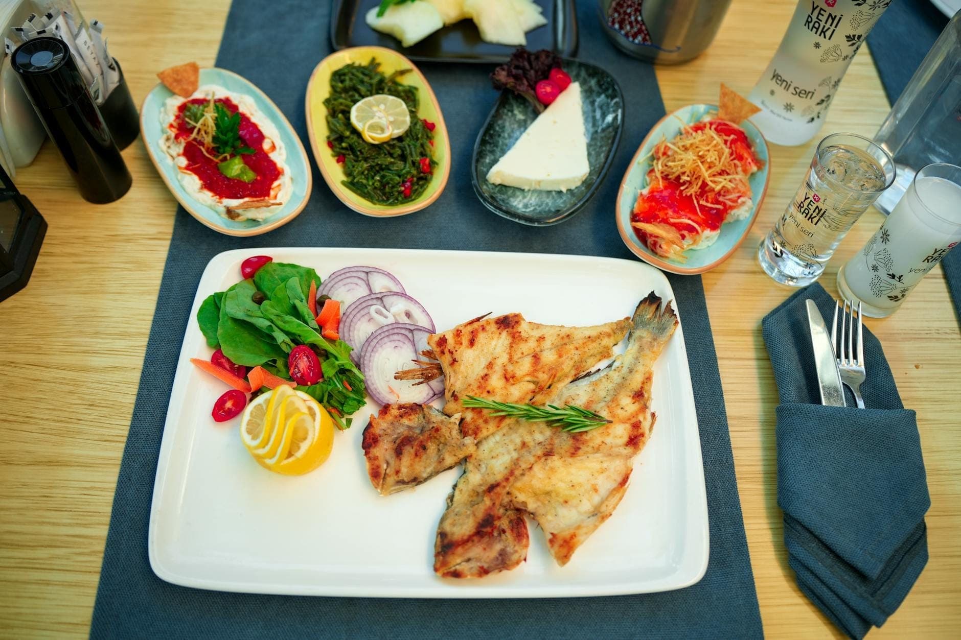 Une table de restaurant bien éclairée avec du poisson grillé, une salade fraîche, des mezes turcs et du raki, mettant en valeur la cuisine méditerranéenne associée à la scène culinaire d'Antalya.