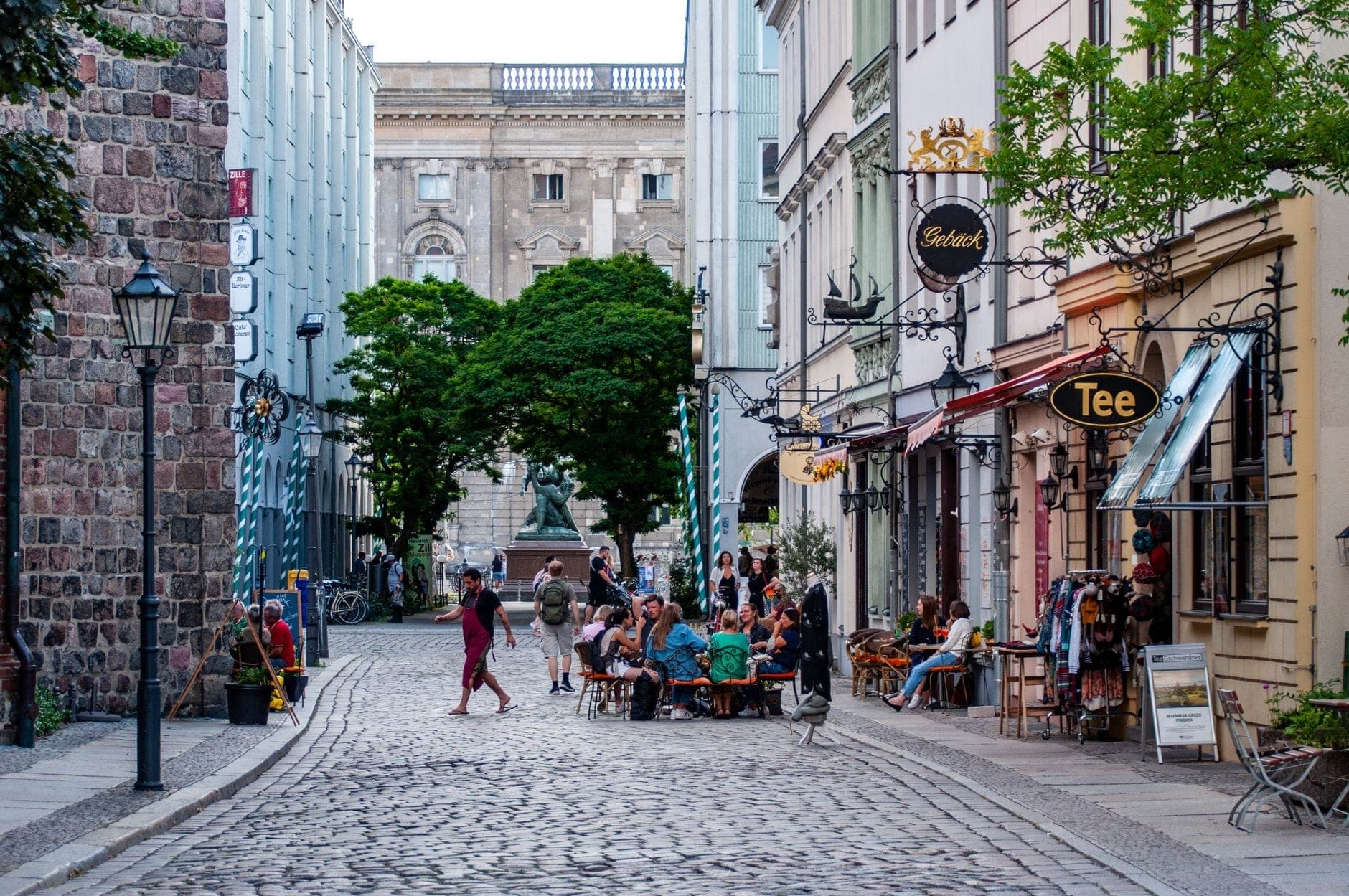 Calle adoquinada en Berlín bordeada de cafés y pequeñas tiendas, personas cenando al aire libre y un edificio histórico al fondo.