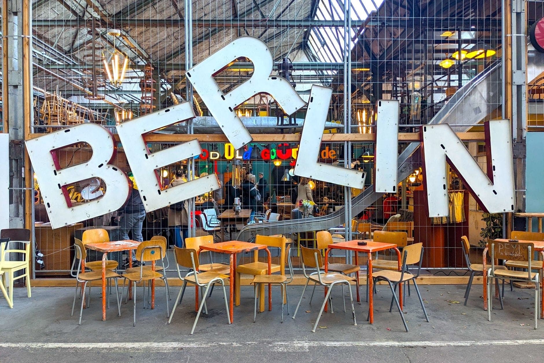 ที่นั่งกลางแจ้งสีสันสดใสในฟู้ดฮอลล์เบอร์ลิน พร้อมป้าย BERLIN ขนาดใหญ่สุดชิค คนกินอาหารนั่งอยู่ด้านในหลังรั้วตาข่าย