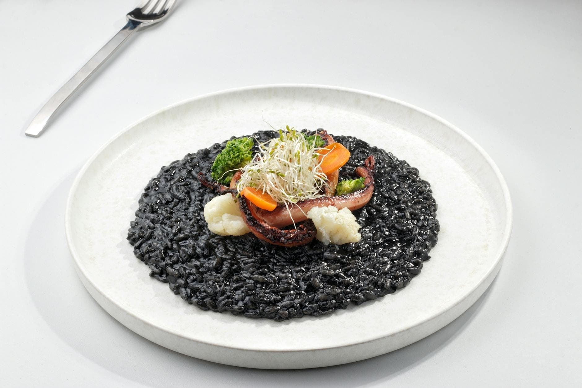 Um prato de risoto negro guarnecido com legumes e brotos, apresentado numa mesa branca com um garfo próximo.