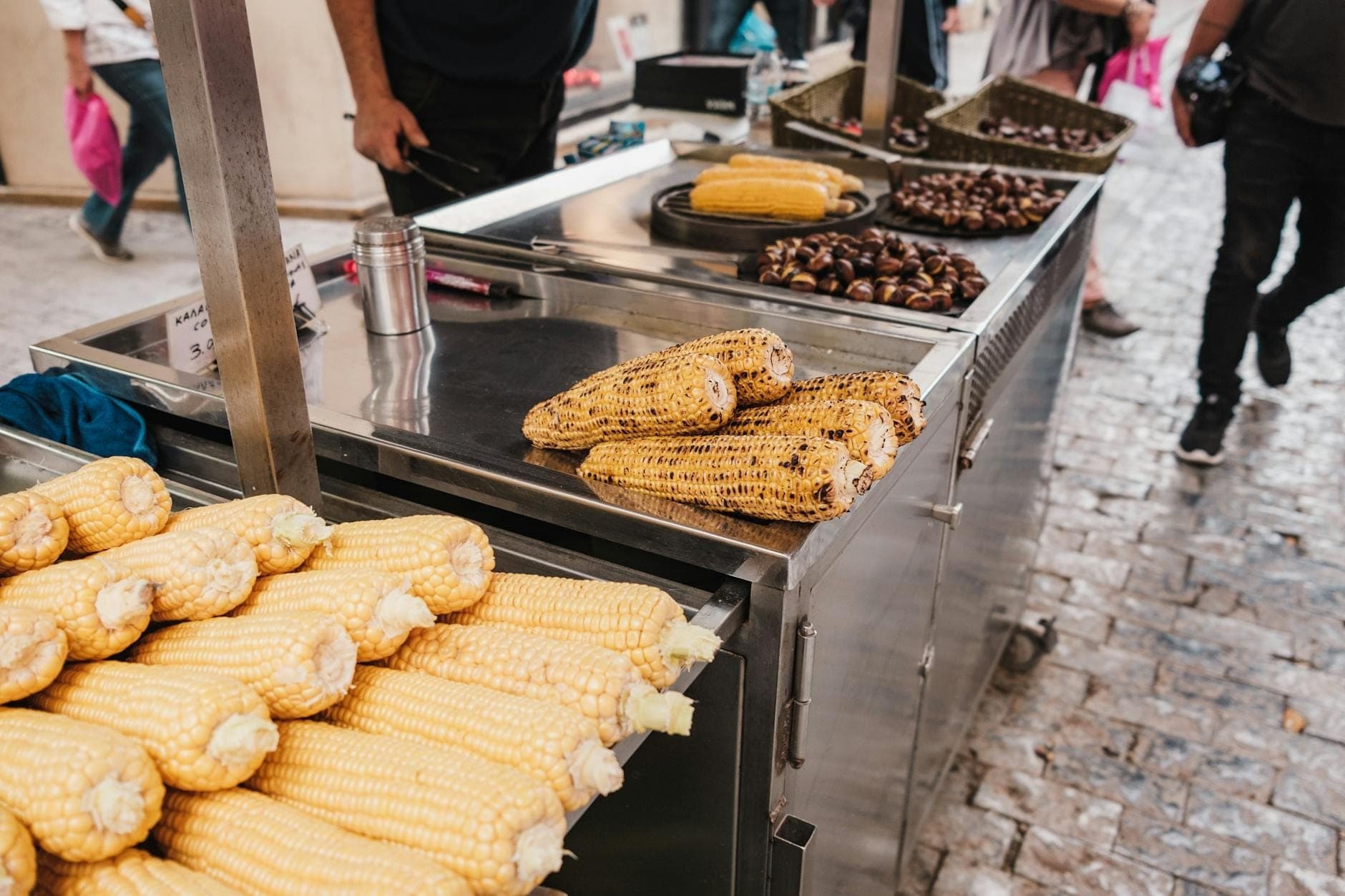 Barraca de comida de rua com milho grelhado e castanhas assadas em uma rua de paralelepípedos, com pessoas caminhando por perto