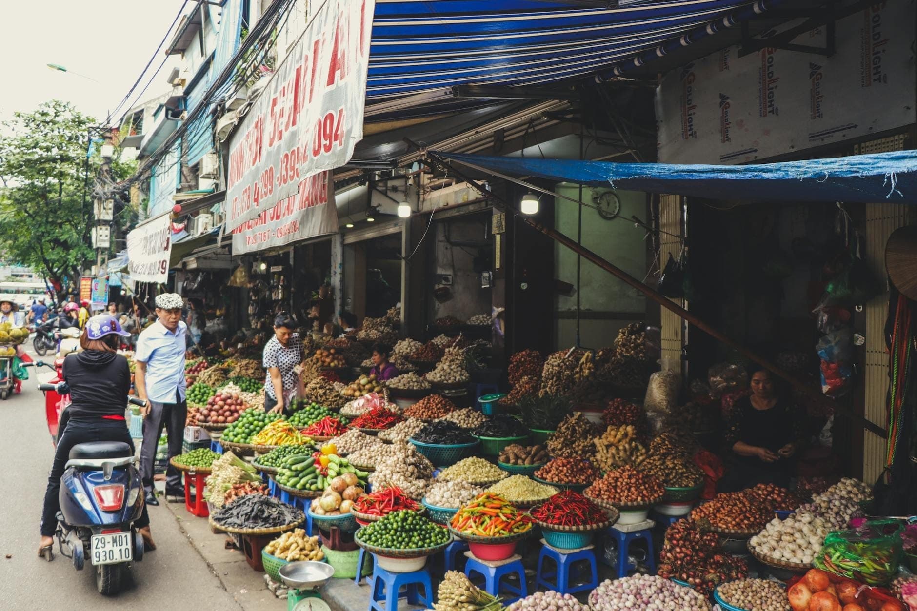 Animado mercado callejero en Hanoi con productos coloridos y vendedores en pequeños taburetes, compradores y scooters pasando junto a frutas y verduras frescas.