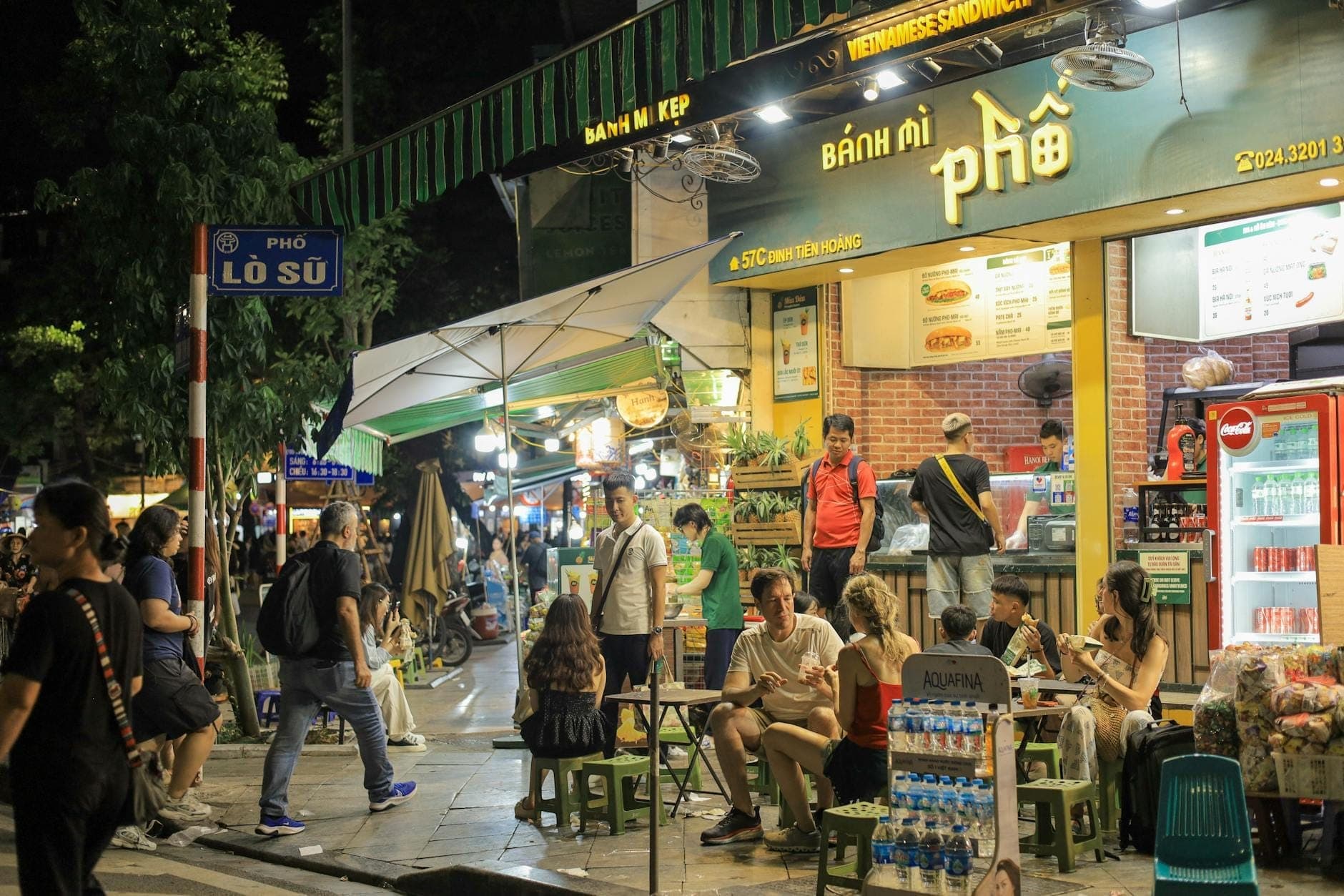 Una animada escena nocturna en una calle de Hanoi con locales y turistas comiendo en mesas al aire libre frente a puestos de comida callejera y restaurantes.