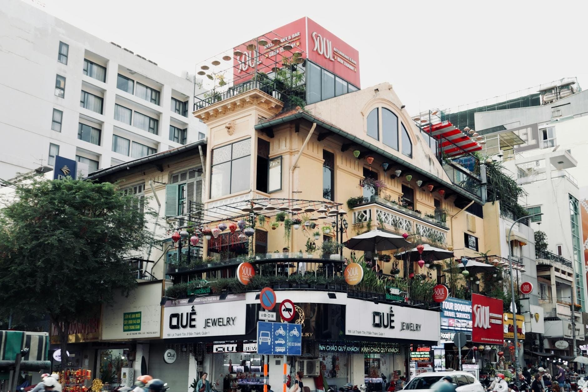 Masiglang sulok ng kalye sa Saigon na may multi-palapag na gusali na may mga restaurant, mga palatandaan sa kalye, mga halaman, at mga taong naglalakad sa downtown ng Ho Chi Minh City.