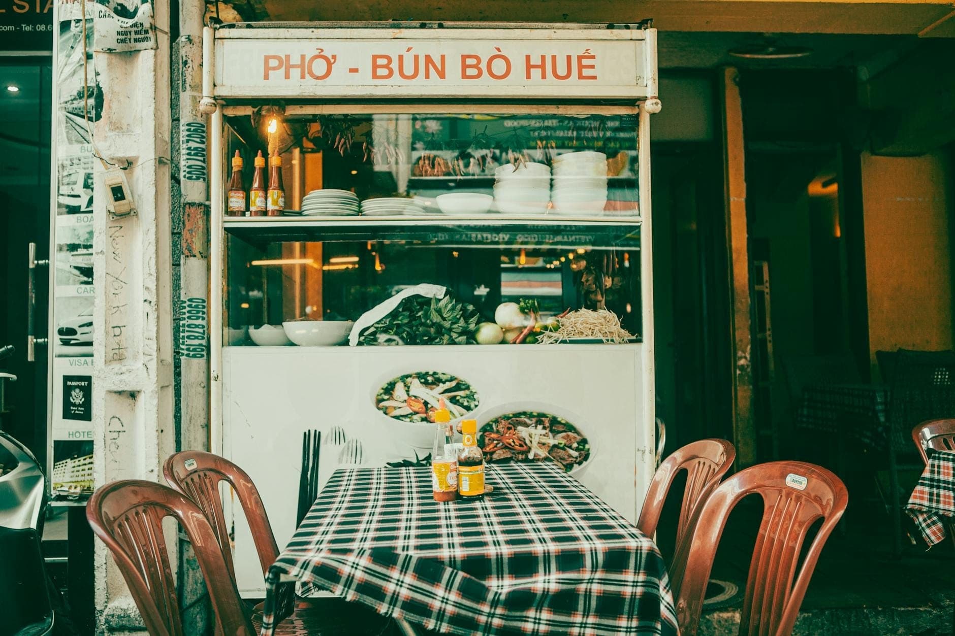 Isang klasikong Vietnamese street food stall na may karatulang 'Phở - Bún Bò Huế', na may mga plastic na upuan at isang mesa na may disenyo ng pabula sa labas.