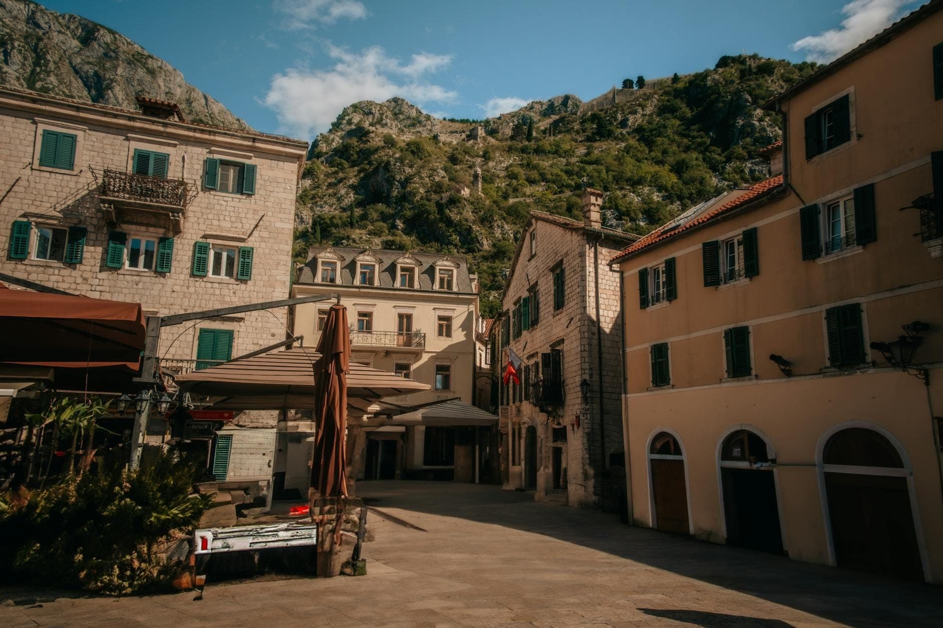 Des bâtiments en pierre aux volets verts entourent une place tranquille dans la vieille ville de Kotor, avec des montagnes se dressant en arrière-plan sous un ciel bleu.