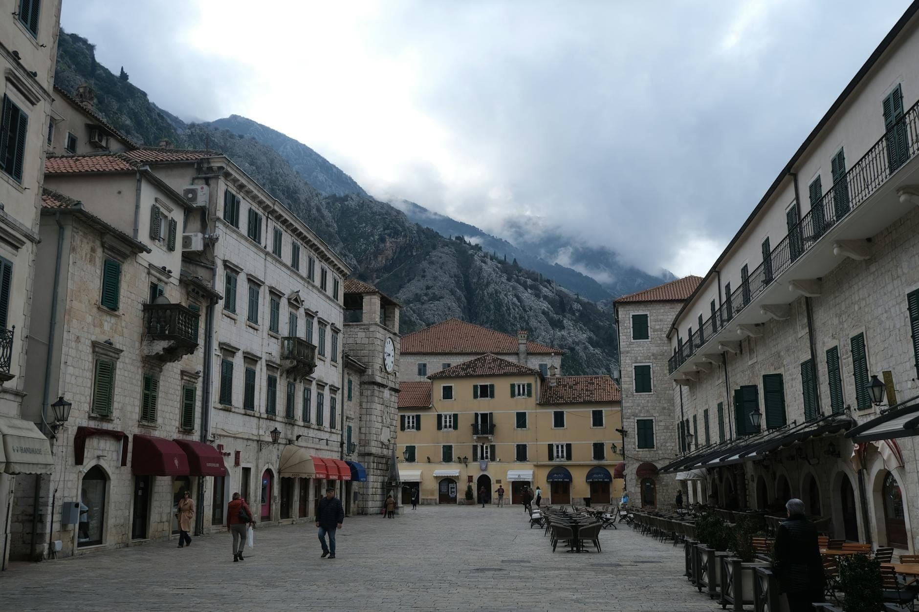 Vaste place pavée dans la vieille ville de Kotor avec terrasse de café, bâtiments historiques, montagnes en arrière-plan et quelques personnes se promenant.