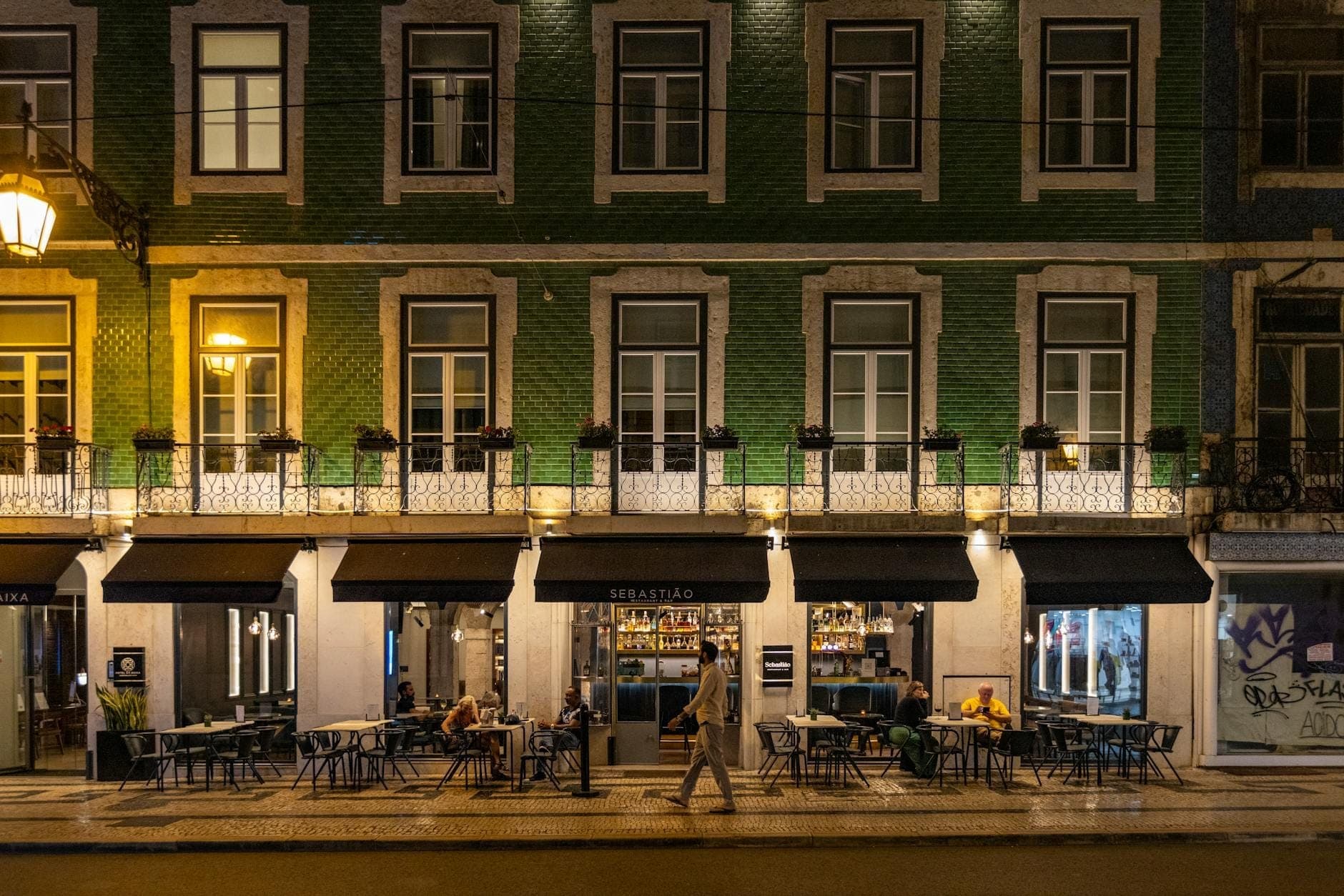 Vista noturna de um elegante restaurante em Lisboa com esplanada, fachada de azulejos verdes e pessoas a jantar numa movimentada rua da cidade.