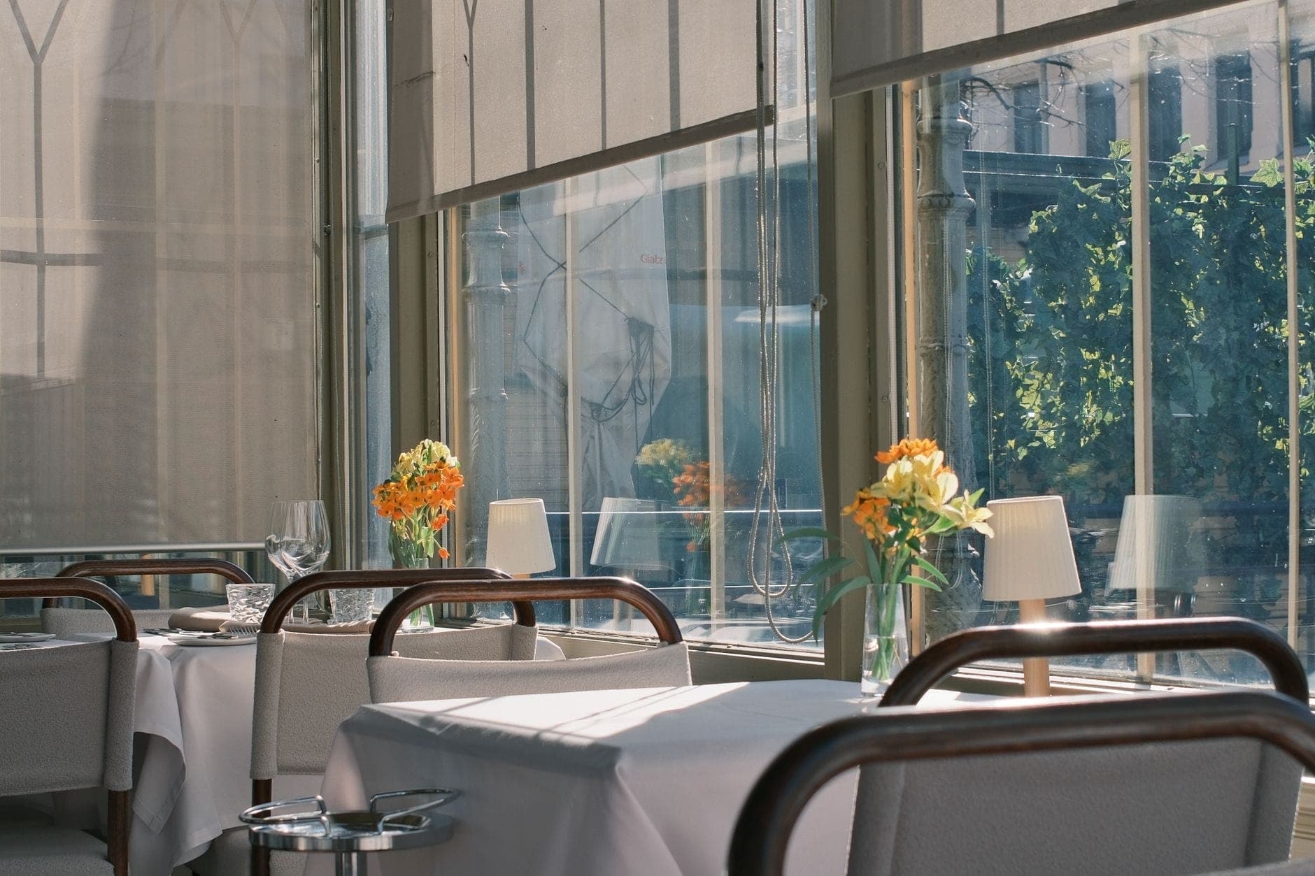 Elegantes Restaurantinterieur bei Tageslicht mit weißen Tischdecken, frischen Blumen und großen Fenstern mit Blick auf eine Stadtstraße.