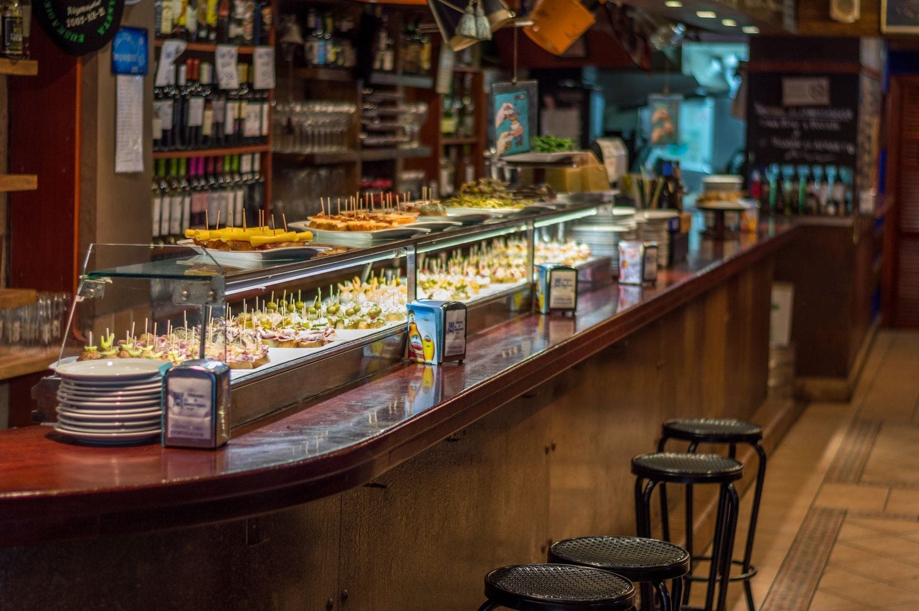 Lange Theke in einem spanischen Tapas-Restaurant mit verschiedenen Pintxos und Tapas auf der Theke, Hockern entlang des Tresens und Weinflaschen dahinter.