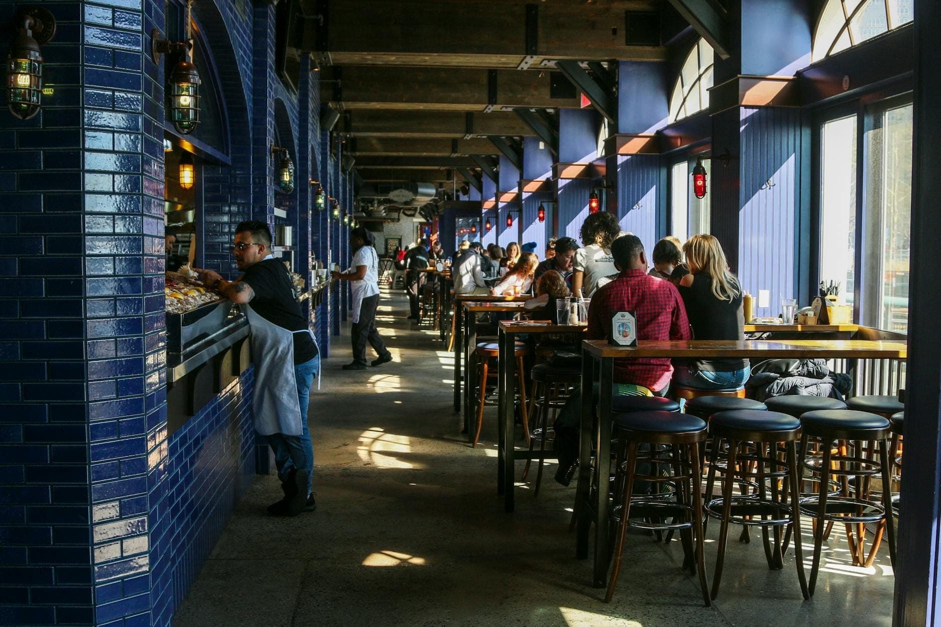 Un restaurant animé et ensoleillé avec des murs carrelés bleus, des clients savourant leurs repas et du personnel servant la nourriture, évoquant la culture culinaire vibrante de New Orleans.