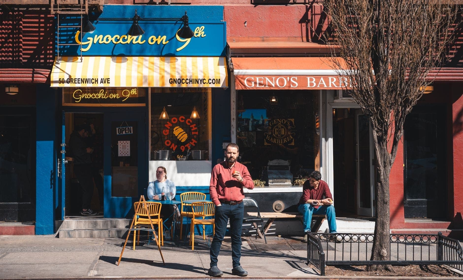 วิวบนถนนของร้านอาหารเล็กสองแห่งใน New York พร้อมที่นั่งกลางแจ้ง หลังคาผ้าสีน้ำเงินและเหลือง คนเดินถนน และบรรยากาศแบบเมือง