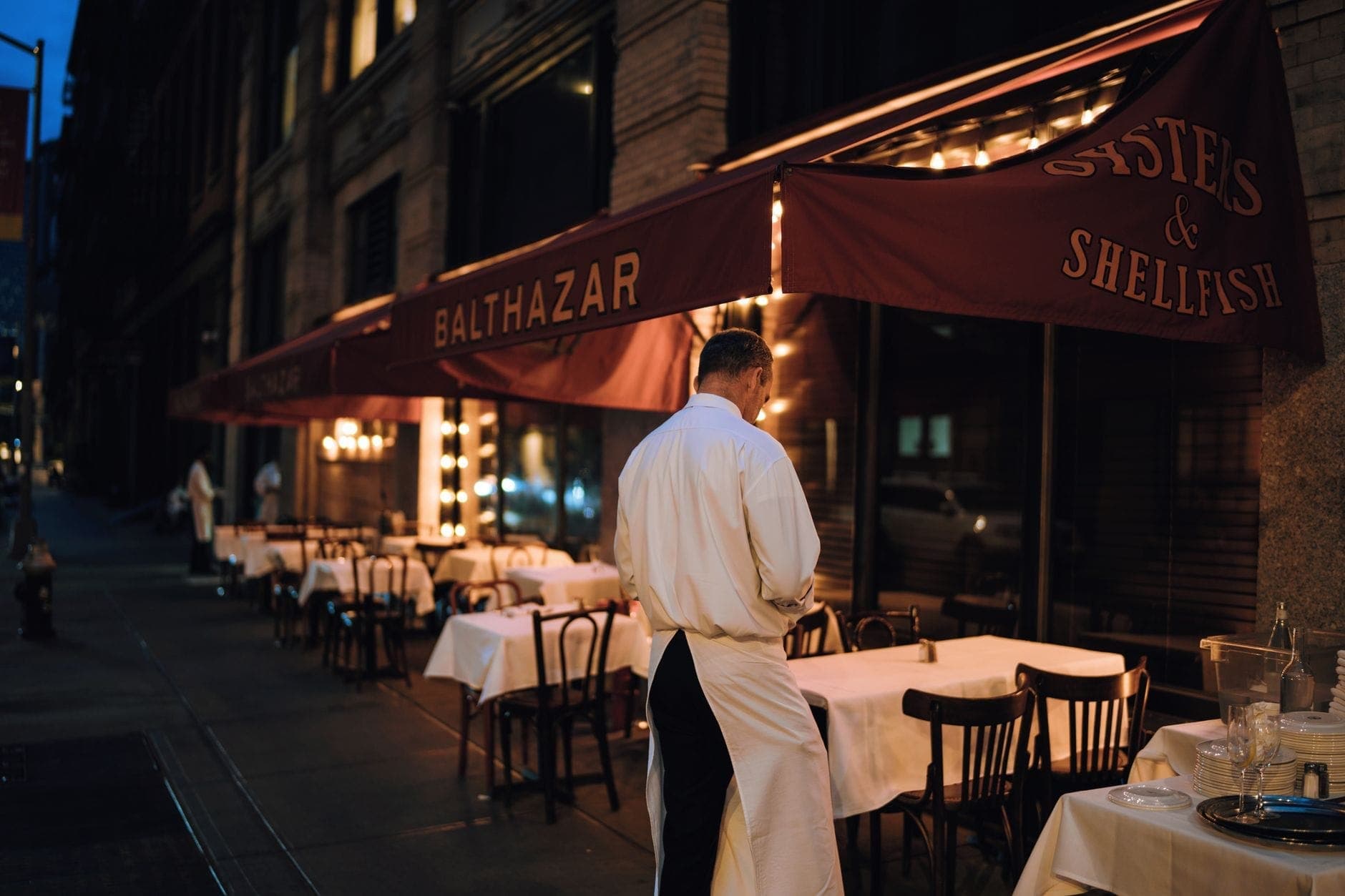 บริกรสวมผ้ากันเปื้อนสีขาวกำลังจัดโต๊ะกลางแจ้งที่ร้าน Balthazar ร้านอาหารไฟน์ไดนิ่งชื่อดังในนิวยอร์กซิตี้ที่มีกันสาดสีแดงในยามเย็น