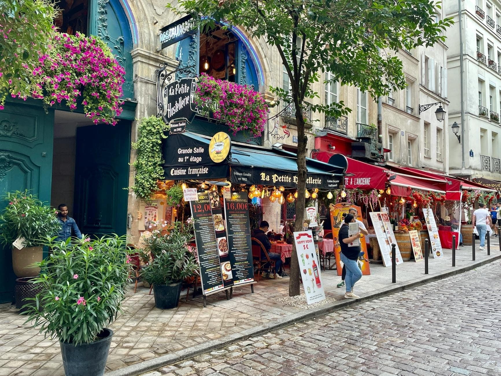 Bustling Parisian street lined with cafes and restaurants, menus and signs outside, people dining and walking, cobblestone pavement.