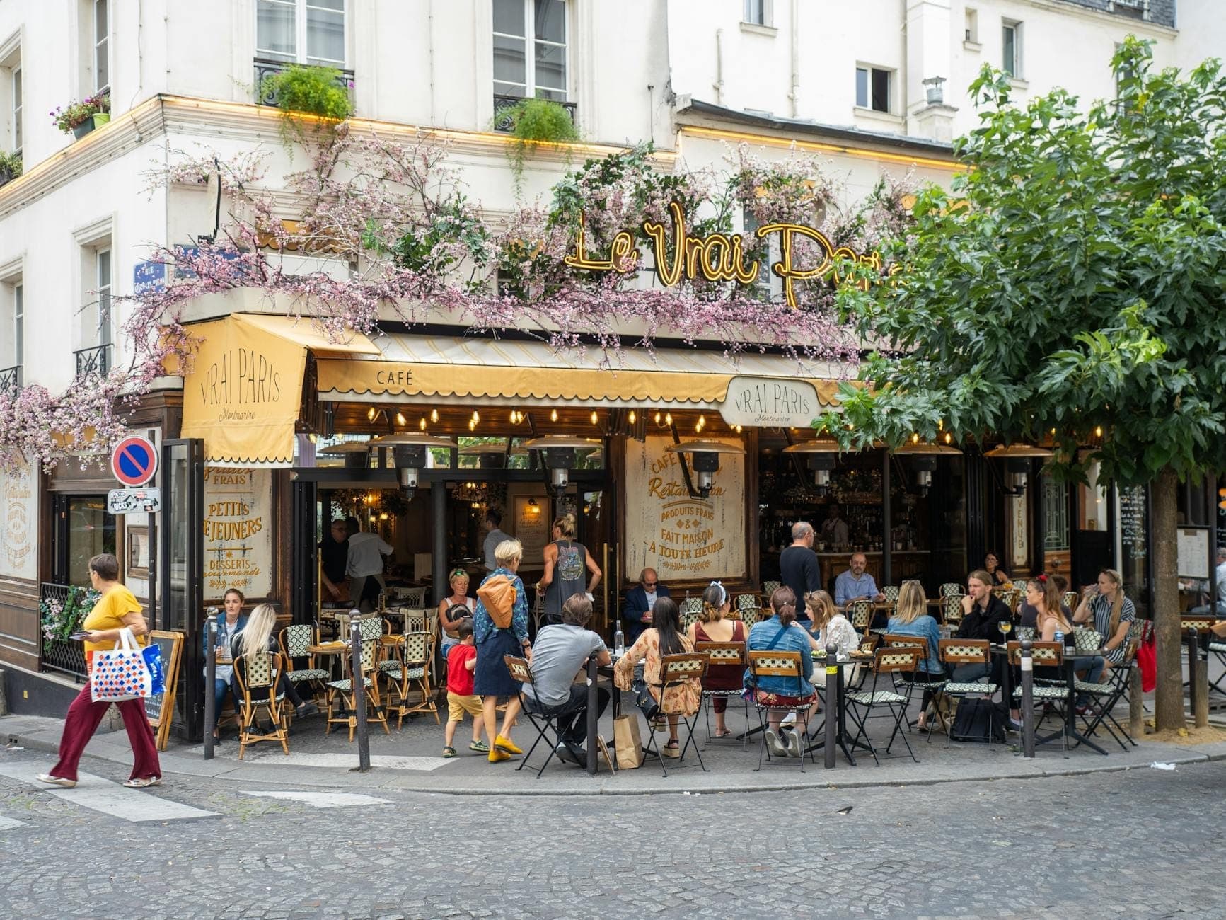 A lively Parisian café with people dining at outdoor tables, flower decorations on the building, and a charming street atmosphere perfect for food lovers.