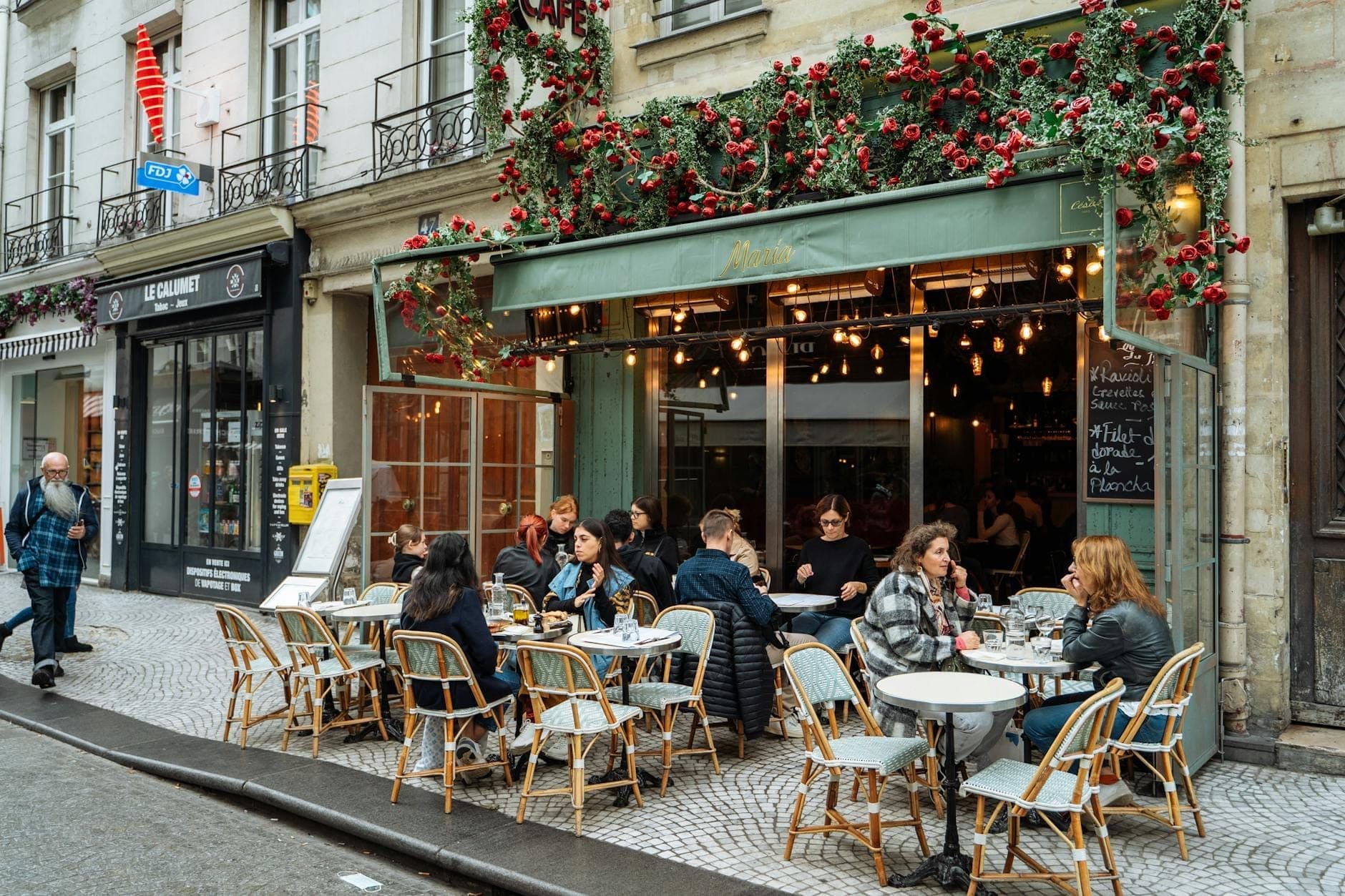 People dining outdoors at a bustling Parisian café with classic chairs and a flower-adorned façade on a city street.