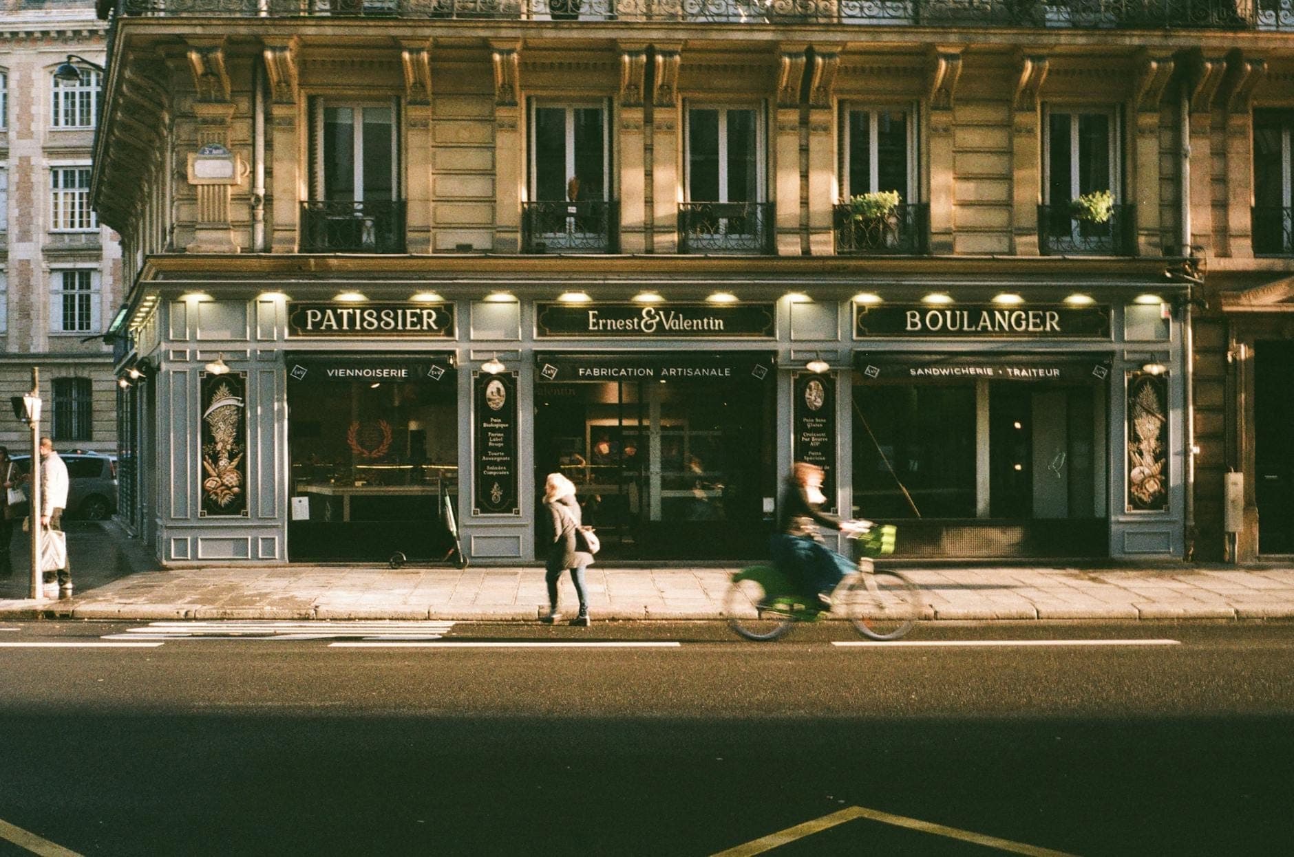 Street view of a Parisian boulangerie and patisserie with people walking and cycling past its classic Paris facade.