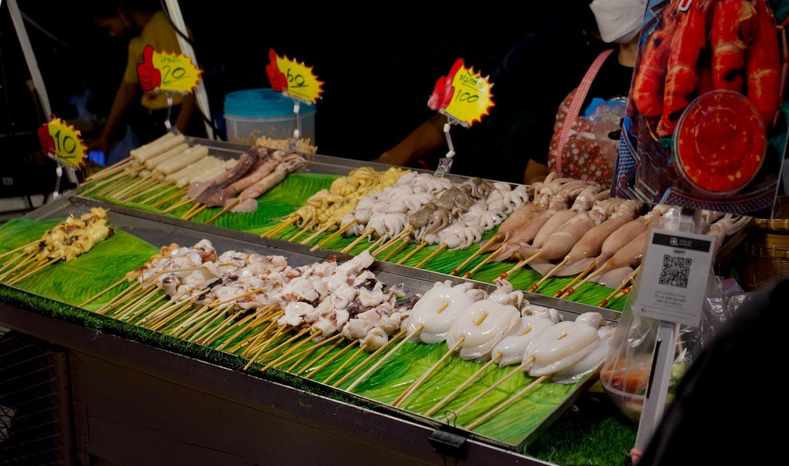 Colorful seafood skewers displayed on a street food stall at night in Pattaya, with prices and vendors visible in the background.