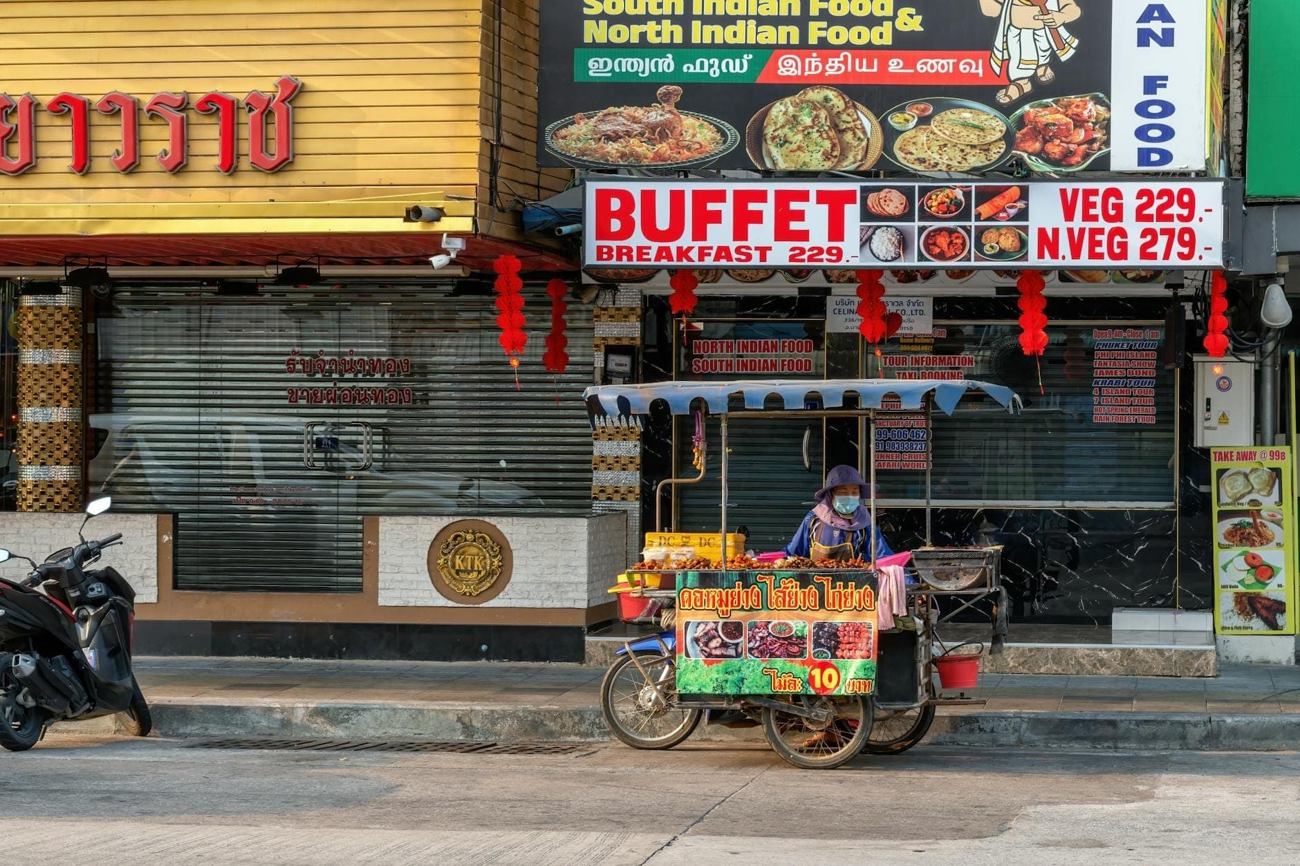 Un carrello di cibo di strada davanti a ristoranti con insegne colorate, tra cui cucina dell'India meridionale, che riflette la variegata scena culinaria di Pattaya.