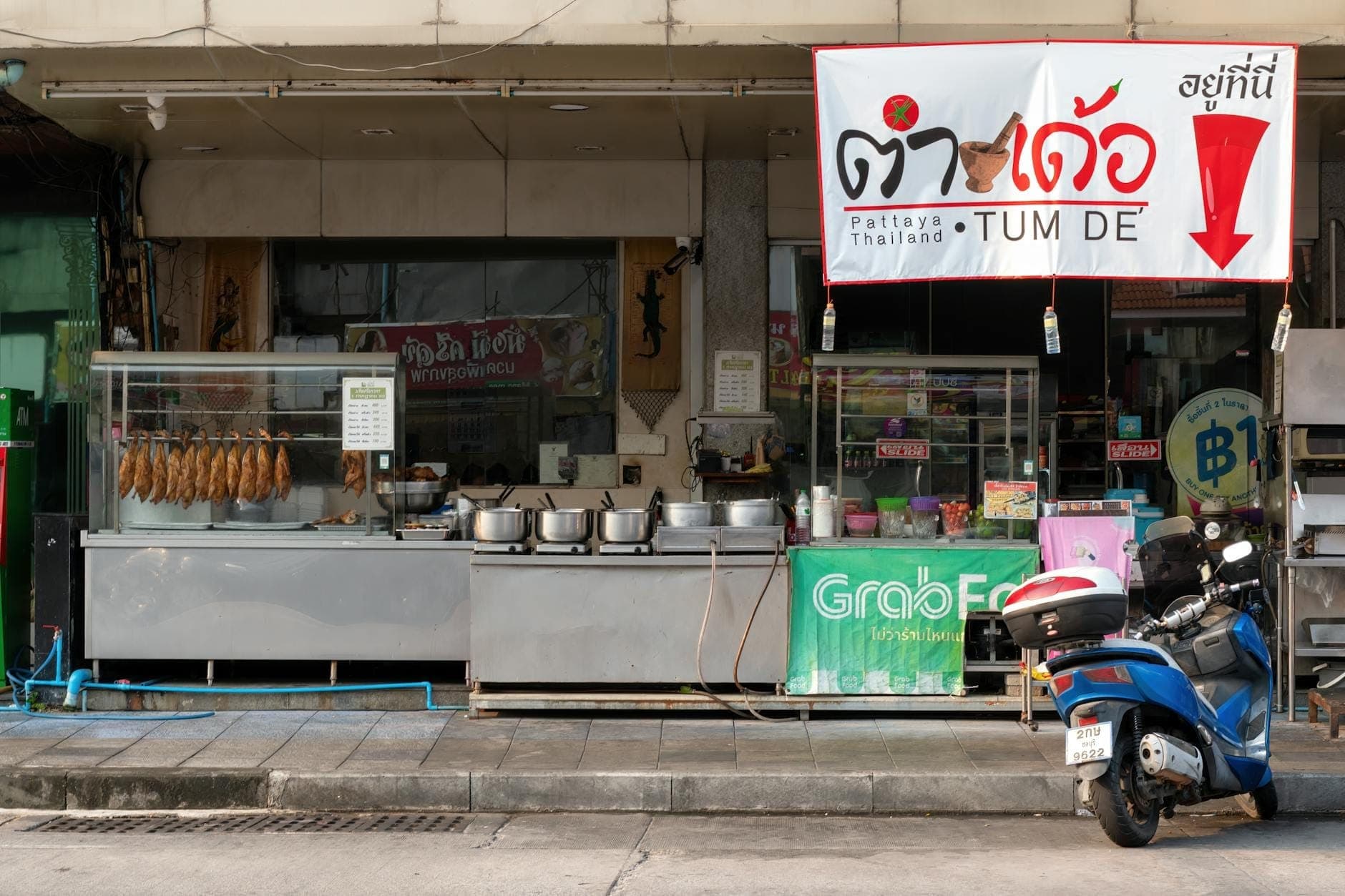 Vista di strada di un tipico ristorante tailandese in stile shophouse a Pattaya, con carretti per il cibo in metallo, pentole, carni appese, scooter e insegne in tailandese.