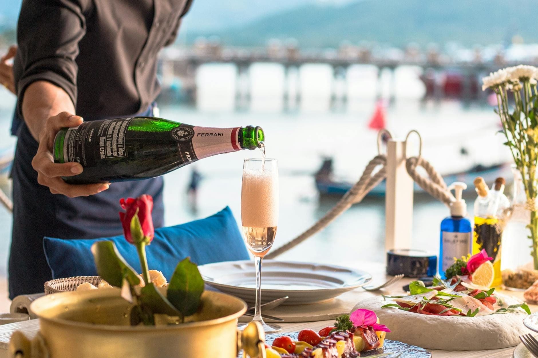 Eleganteng hapag-kainan sa tabi ng dagat na may magagandang pagkain at champagne na ibinubuhos, na may tanaw sa isang pantalan at tubig sa Phuket.